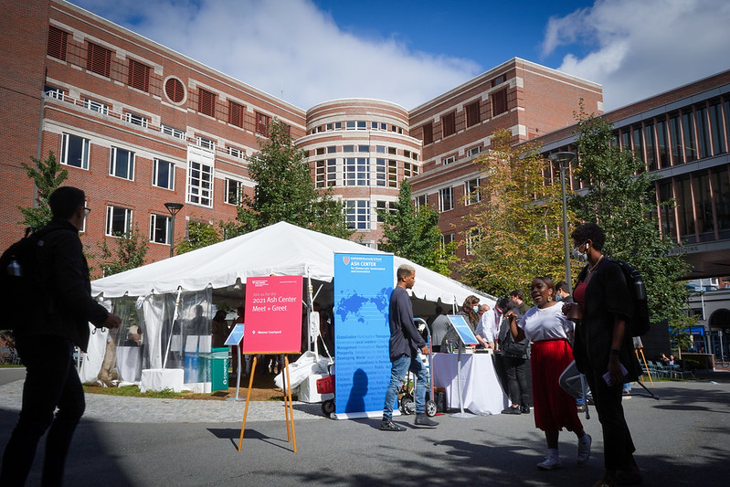 A white tent sits in the middle of Wexner Courtyard surronded by brick and glass Harvard Kennedy School buildings, people are moving in and out of the tent while students walk by on the walkways crisscrossing the courtyard