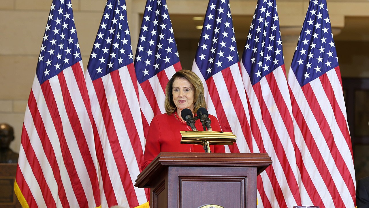 Speaker Pelosi, wearing a red suit, stands behind a podium, behind her is a row of tall American flags