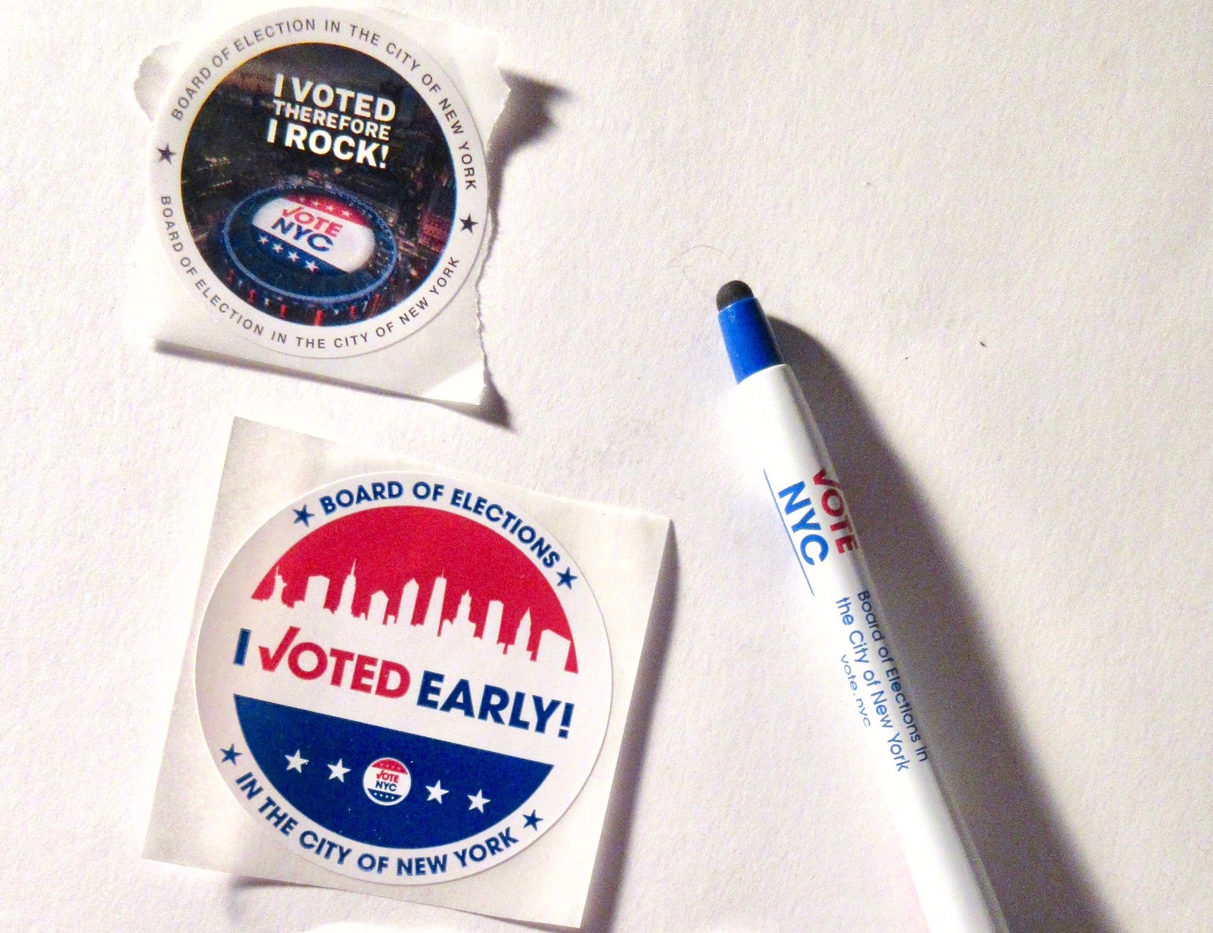 A sticker reads "Board of Election in the City of New York, I voted therefore I rock!" and another reads "I voted early." Next to the stickers is a red, white, and blue pen that reads "Vote NYC, Board of Elections in the City of New York"