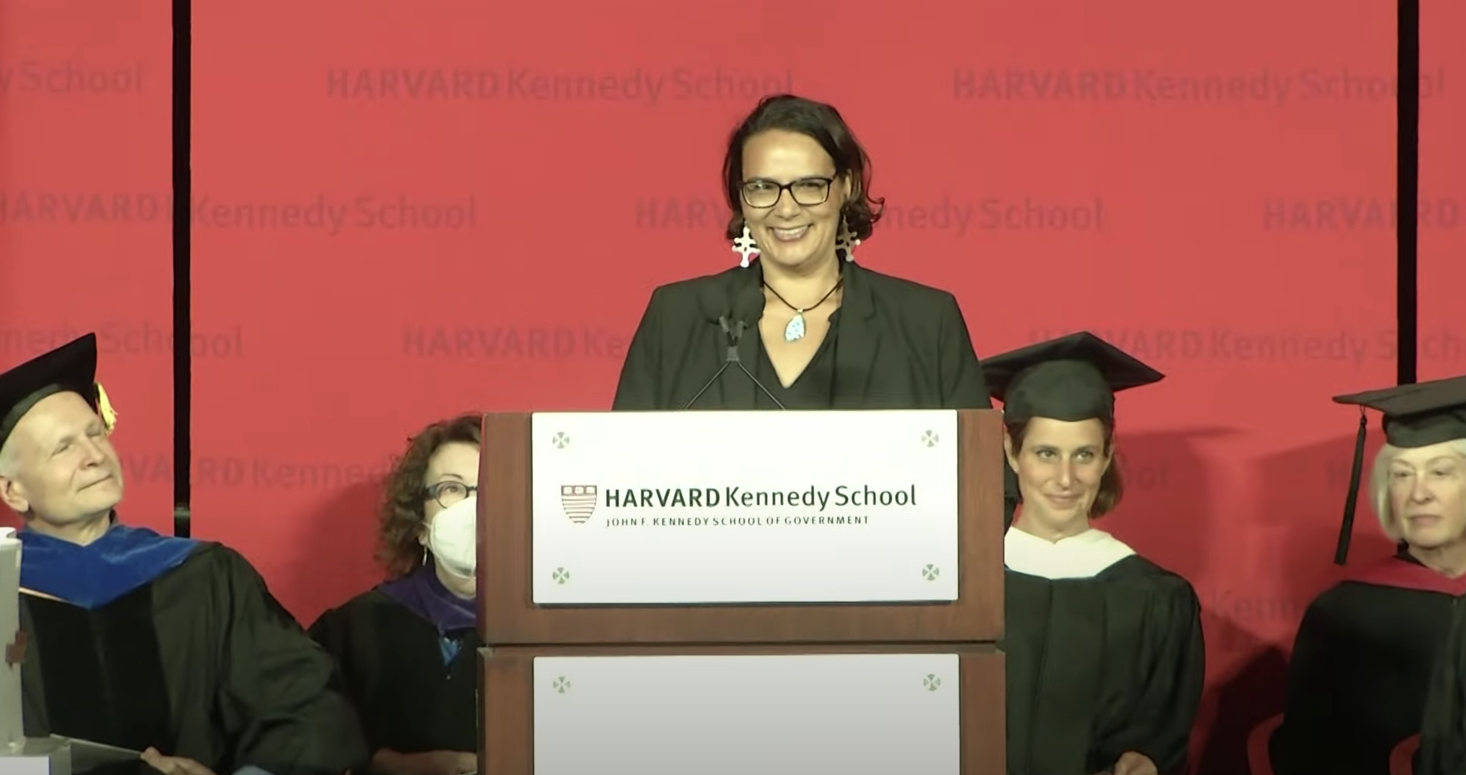 Megan Minoka Hill stands at a podium addressing Kennedy School graduates