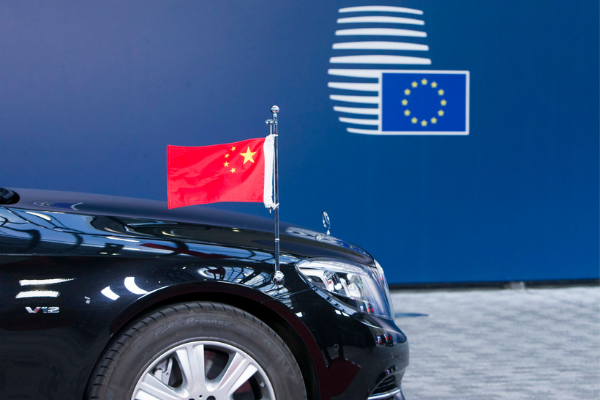 A Chinese flag flies on the hood of a car, in the background the flag of the European Union is seen on a blue wall