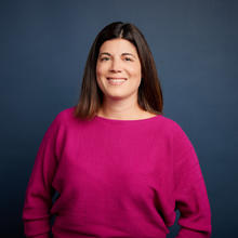 Melissa stands against a dark blue background. She is smiling and wearing a fuchsia sweater.