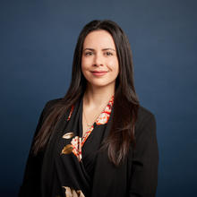 Sarah stands against a dark blue background. She is smiling and wearing a black blazer with a black blouse underneath with a orange and red flower pattern.