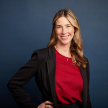 Caryn stands smiling against a dark blue background, she wears a black tailored suit jacket and a bright red blouse