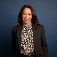 Sadie stands in front of a dark blue background. She is smiling with her hands in her pockets. She's wearing a black blazer with a black, white, and pink flowered blouse.