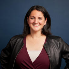 Miriam stands against a dark blue background with her hands on her hips. She is wearing a maroon sweater and a leather jacket.