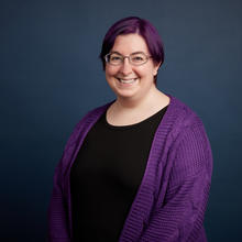 Holly stands in front of a dark blue background, they are wearing a purple sweater that matches their purple hair